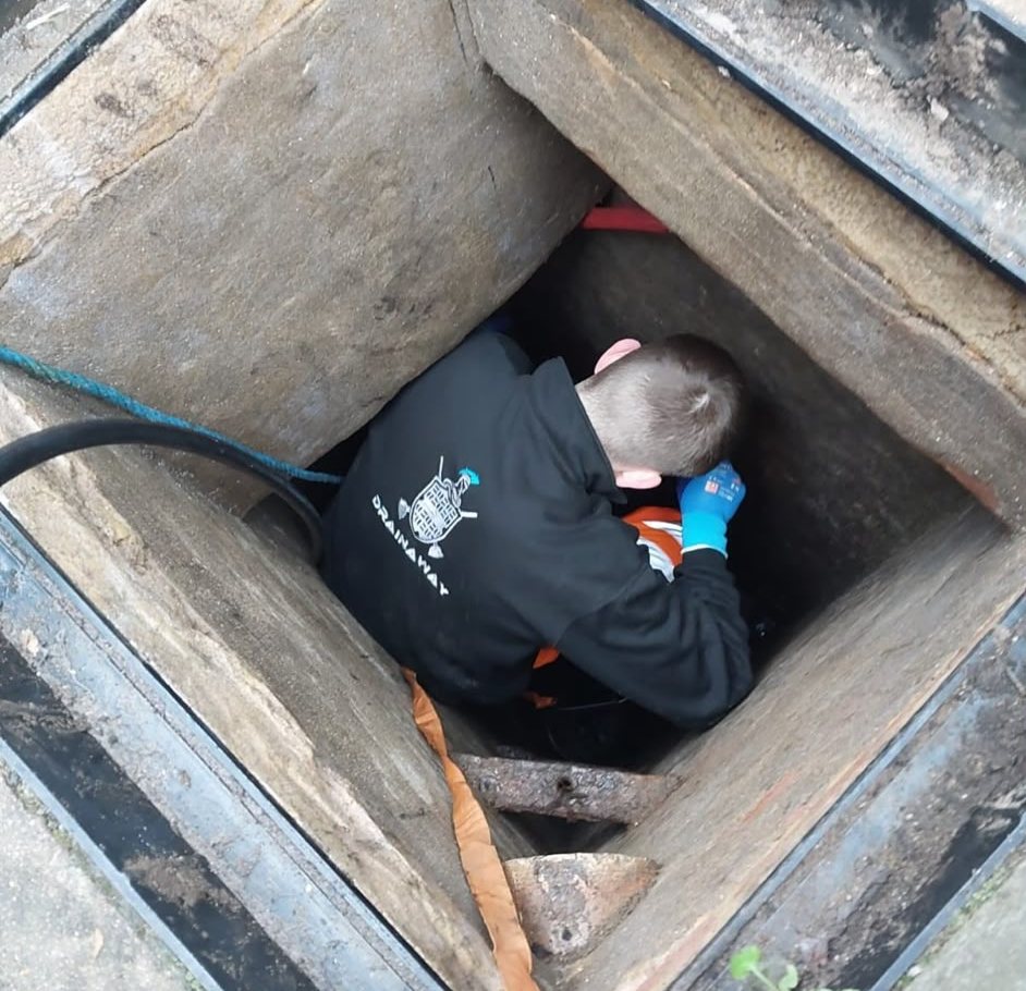 A young apprentice in a DrainAway hoodie leaning down inside an open inspection chamber to reach the pipe