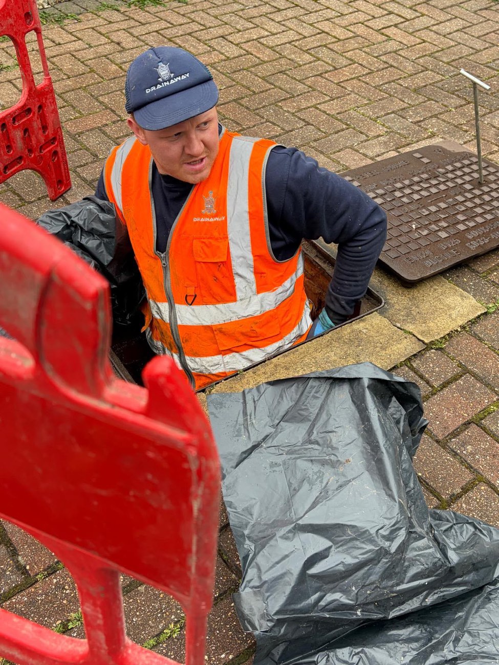 A DrainAway engineer in a high-visibility vest working in an open manhole behind safety barriers