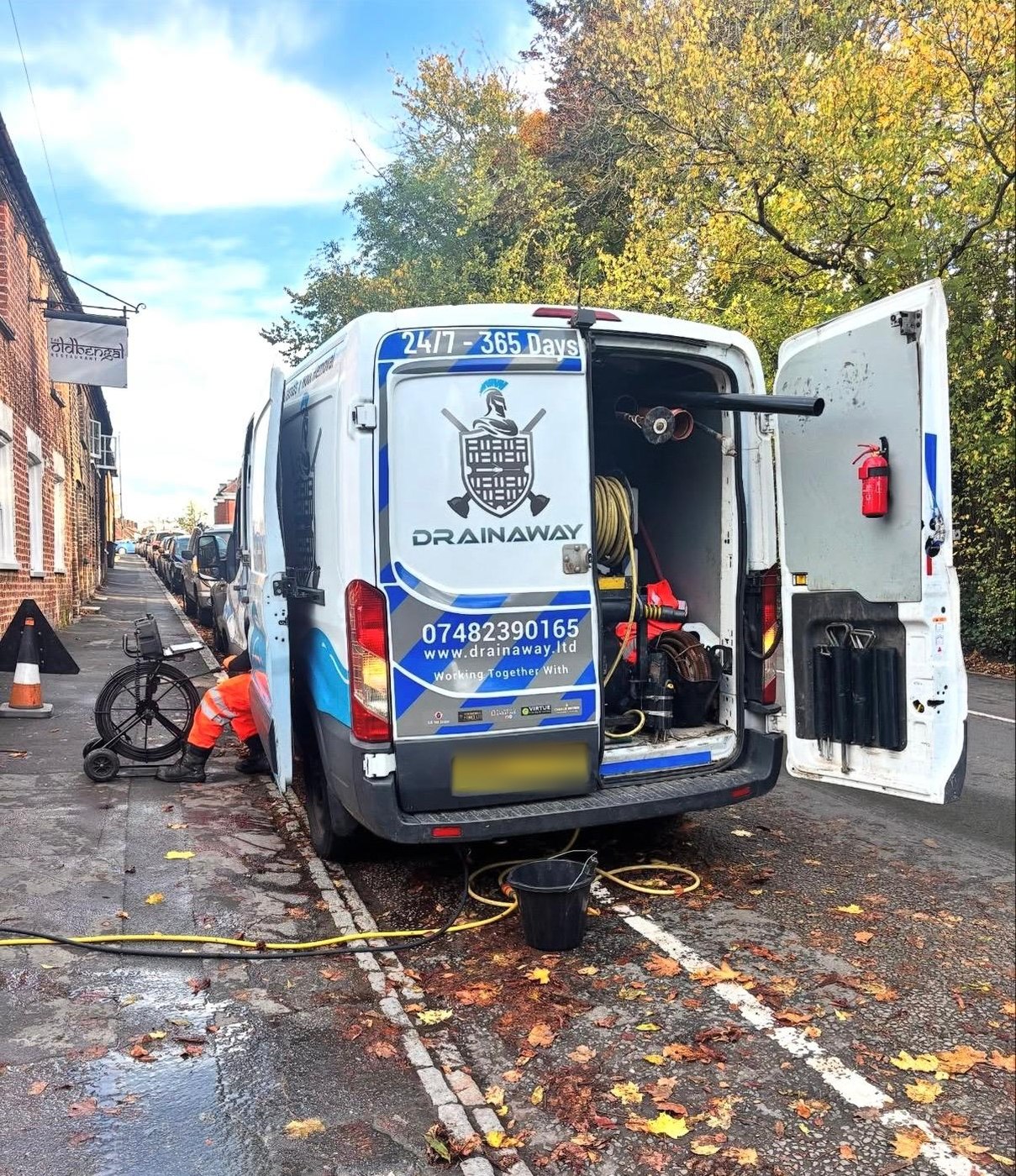 A DrainAway van parked on a street with its rear doors open and the jetting gear out, an engineer working at a drain alongside it