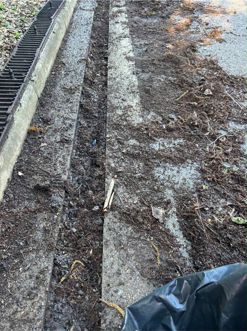 A car park channel drain packed solid with silt, mud and leaf debris, its guards lifted out along the length of the run.
