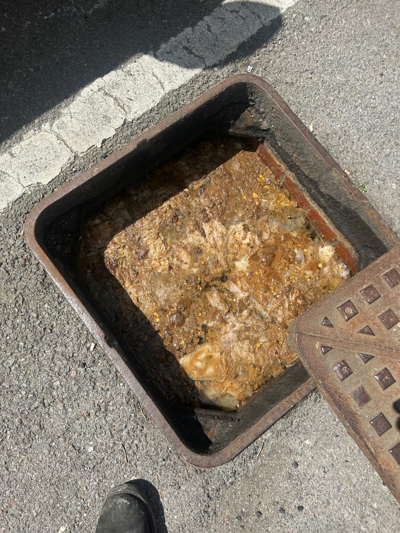 A roadside inspection chamber filled to the top with congealed fat, grease and food waste, the cover lifted off beside it.