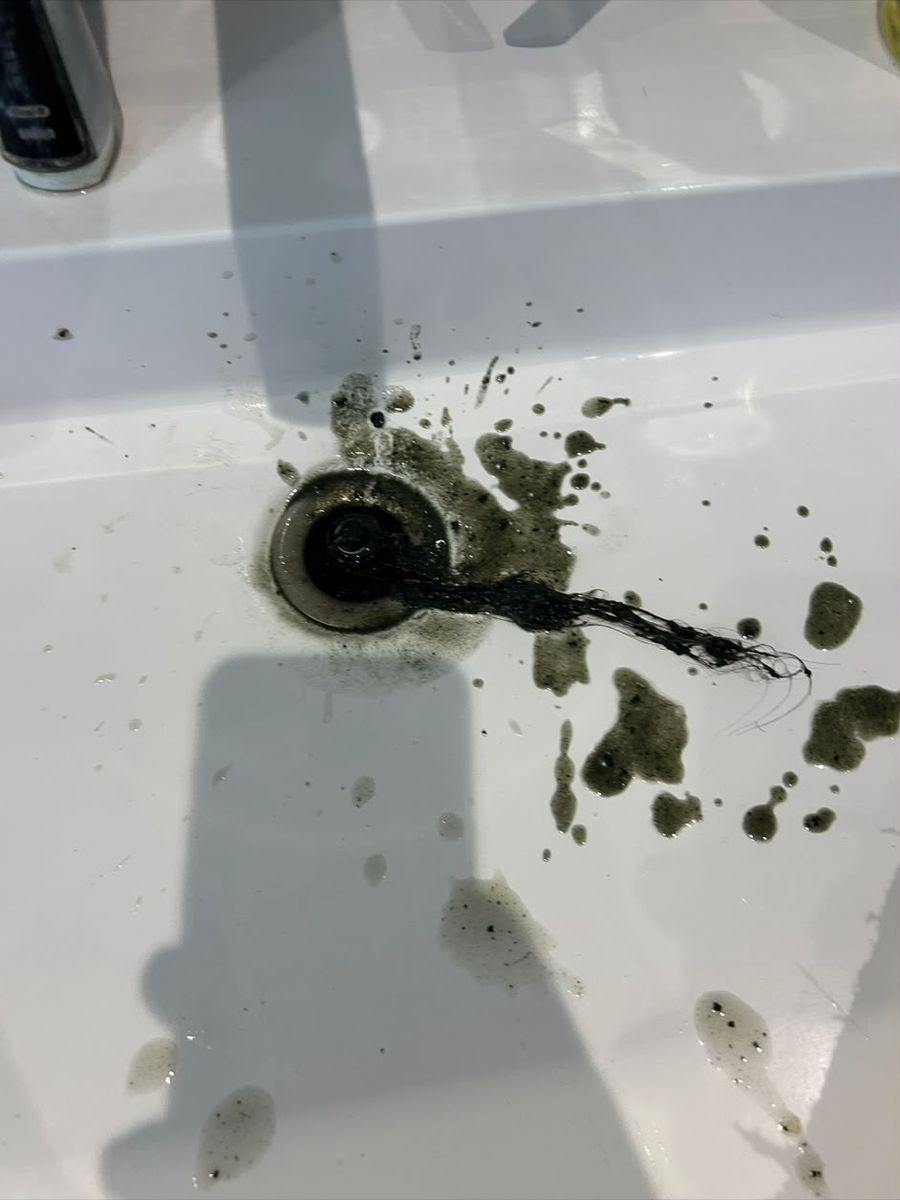 Black silt and a clump of hair pulled up out of a bathroom basin waste and spread across the white basin around the plughole.