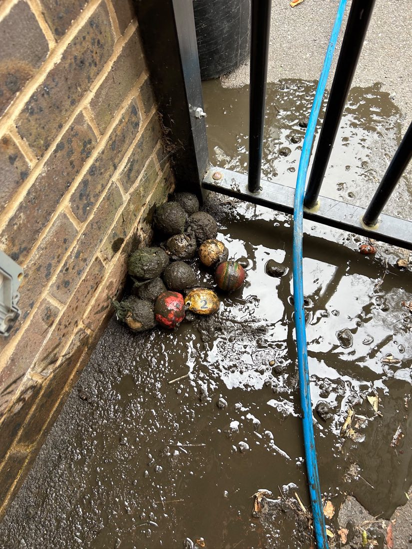 Muddy playground balls recovered from a blocked drain, piled against the wall of a flooded school playground with a jetting hose running across it.