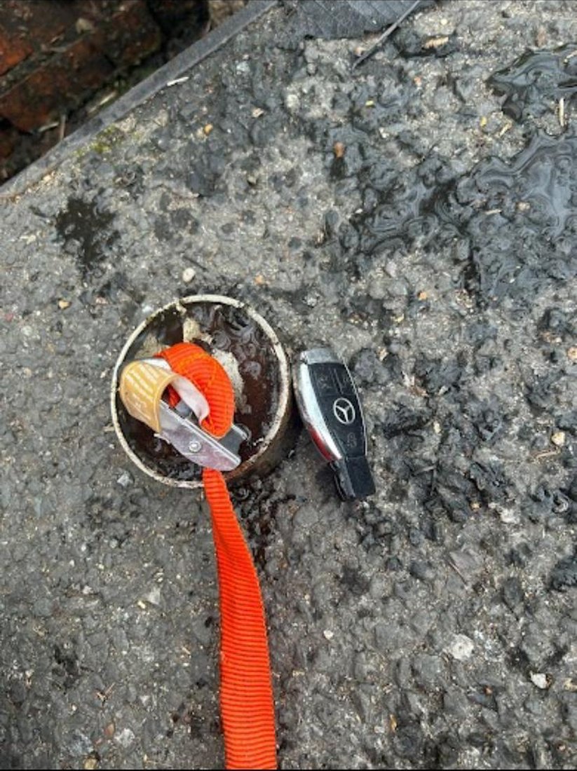 The recovered car key laid on the tarmac beside the open gully pot, with the retrieval tool still on its strap.