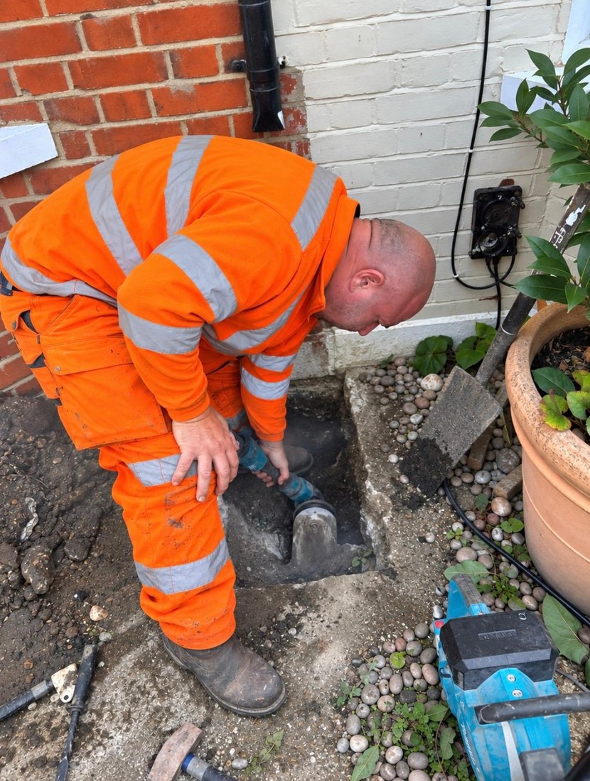 A DrainAway engineer bent over an open inspection chamber set into a gravelled side passage, with a power breaker on the ground beside him.