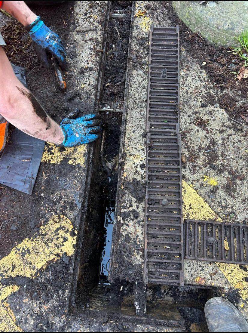 Gloved hands clearing silt and debris out of an open channel drain, with the cast iron grating lifted off alongside it.