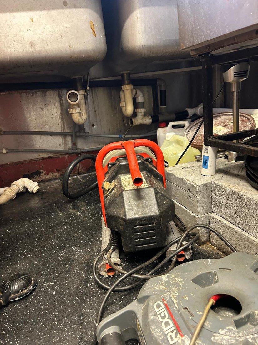 A drain machine set up under the sinks in a commercial kitchen, with the waste pipework opened up above it.