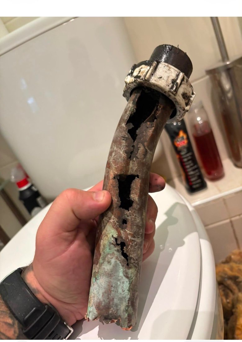 A cut-out length of copper waste pipe held in a gloved hand, rotted through in three places along its length.