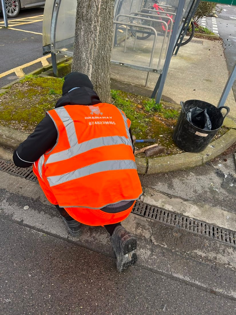 A DrainAway engineer in a high-visibility vest clearing a blocked channel drain running along the kerb.