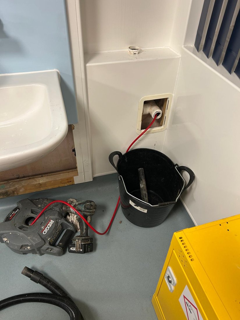 A RIDGID flex shaft machine on a washroom floor, its red shaft fed through a wall access point into the waste pipe above a bucket.
