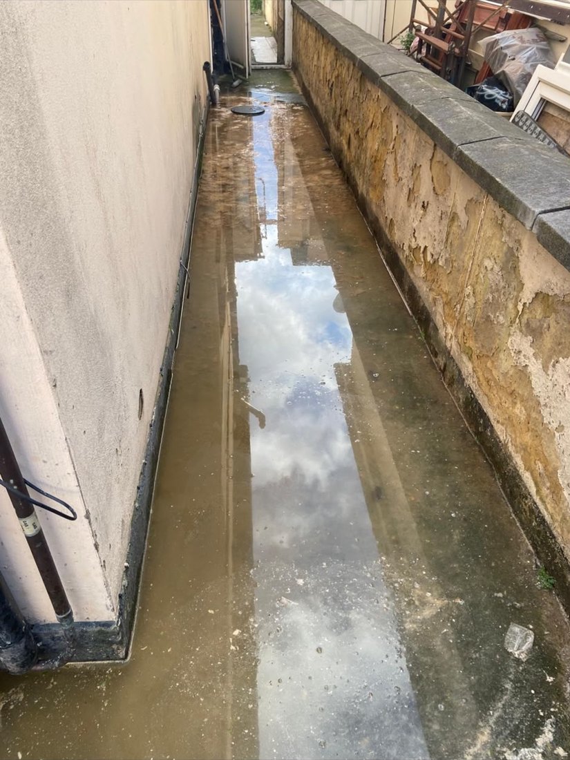 A narrow alley between two buildings standing under dirty water, the sky reflected in it and the gully at the far end submerged.