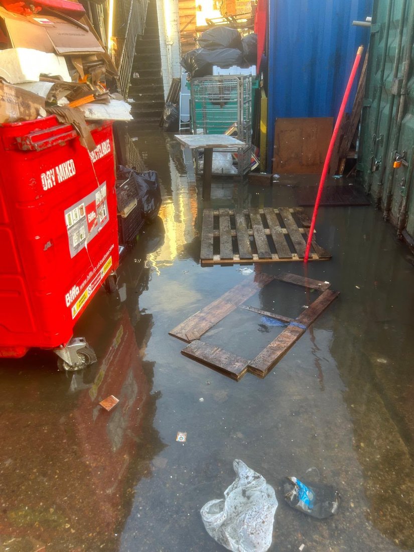 A commercial service yard flooded ankle deep, with a waste bin, pallets and a jetting lance stood in the standing water.