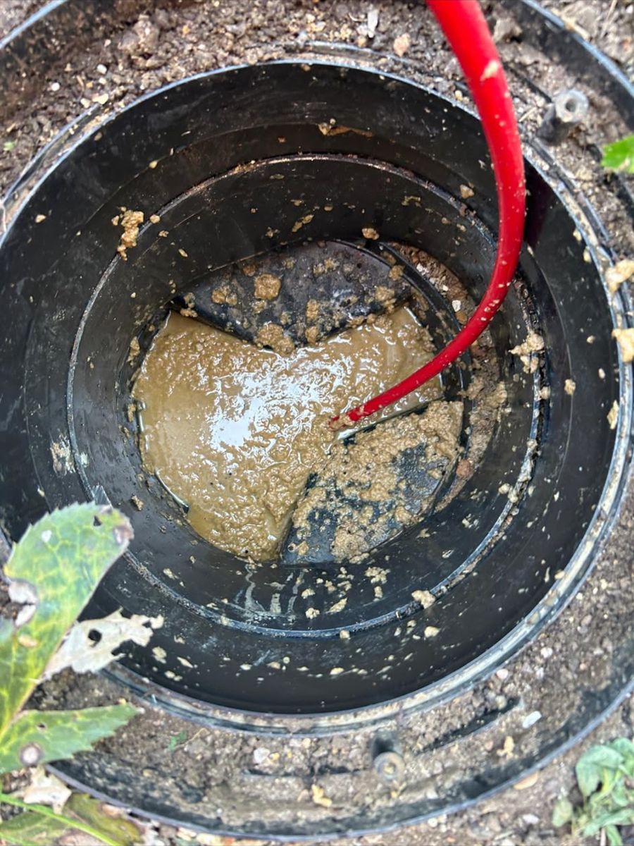 A round inspection chamber set in soil, its base thick with fat and scum, with a red jetting hose fed down into it.