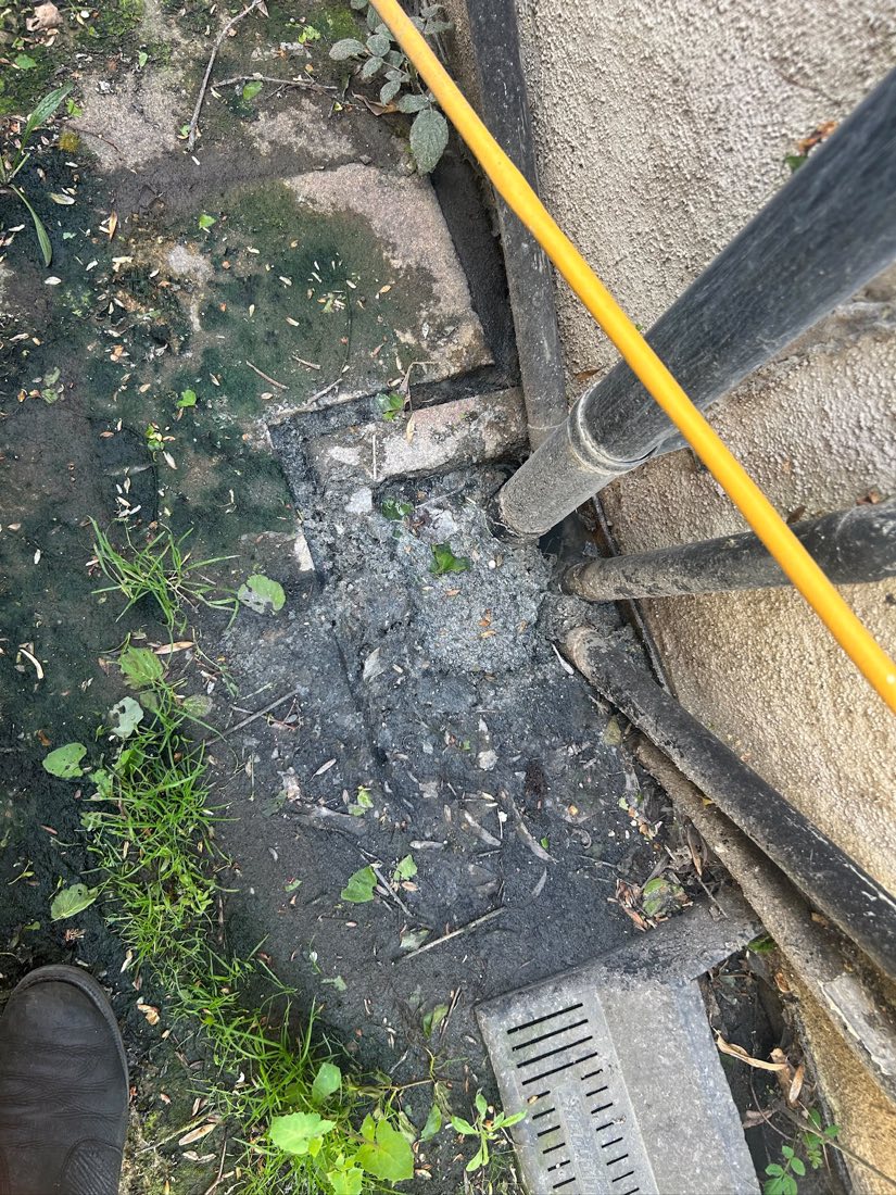 A jetting hose fed into a blocked gully beside a wall, the channel around it thick with silt and leaf debris.