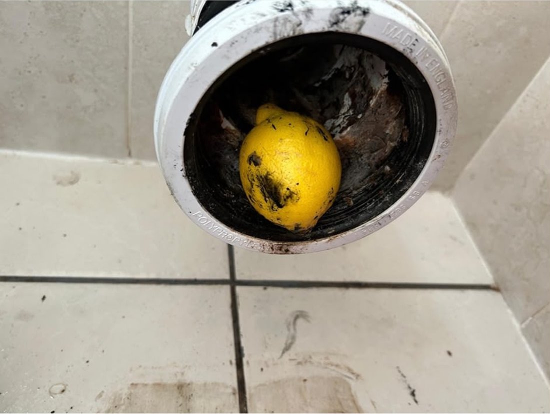 A whole lemon lodged in the mouth of a white waste pipe, held over a tiled bathroom floor.
