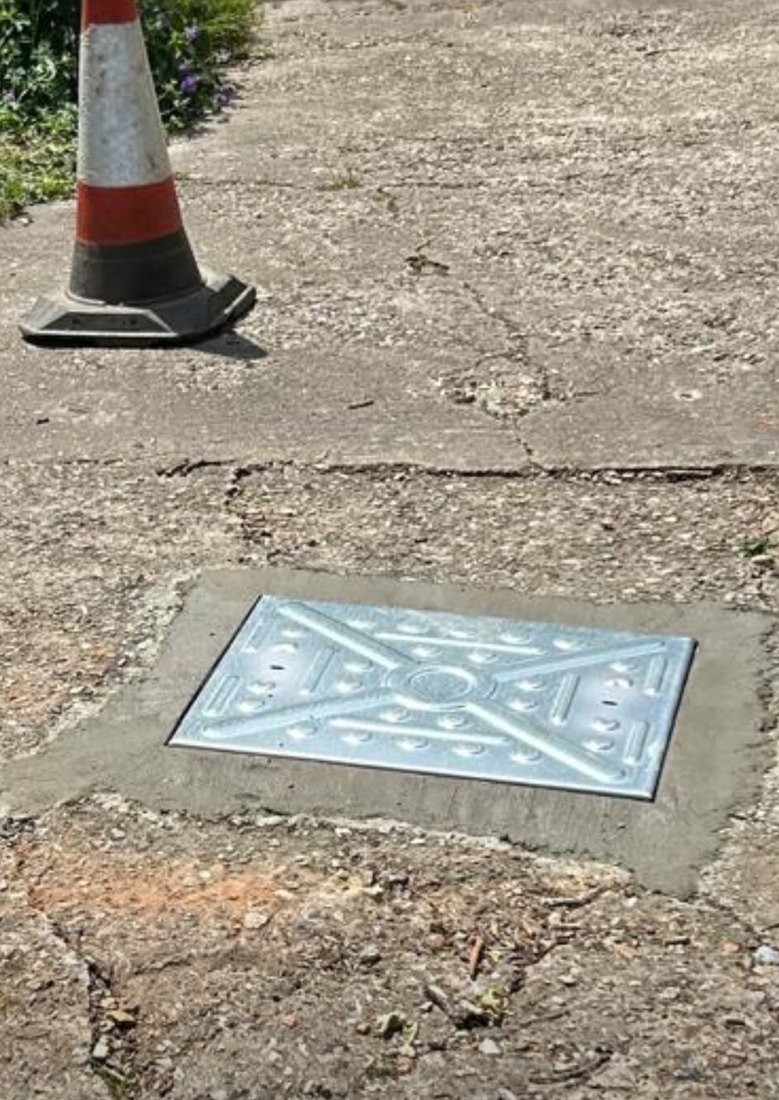 A new galvanised manhole cover bedded into fresh concrete in a driveway, with a cone set beside it.
