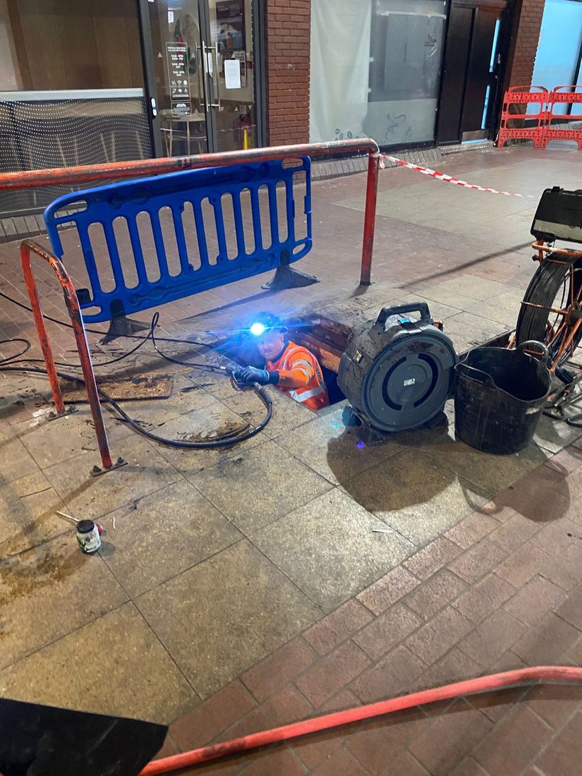 A DrainAway engineer down an open chamber at night in a pedestrianised precinct, working a jetting hose with barriers and tape set out around the manhole.