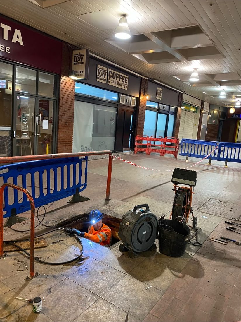 A DrainAway engineer down an open chamber at night in a covered shopping parade, working by head torch with barriers and a jetting reel set around the opening.