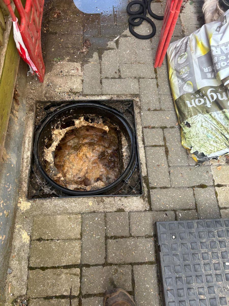 An inspection chamber set in block paving, brimming with set fat and food waste, with barriers up around the open cover.
