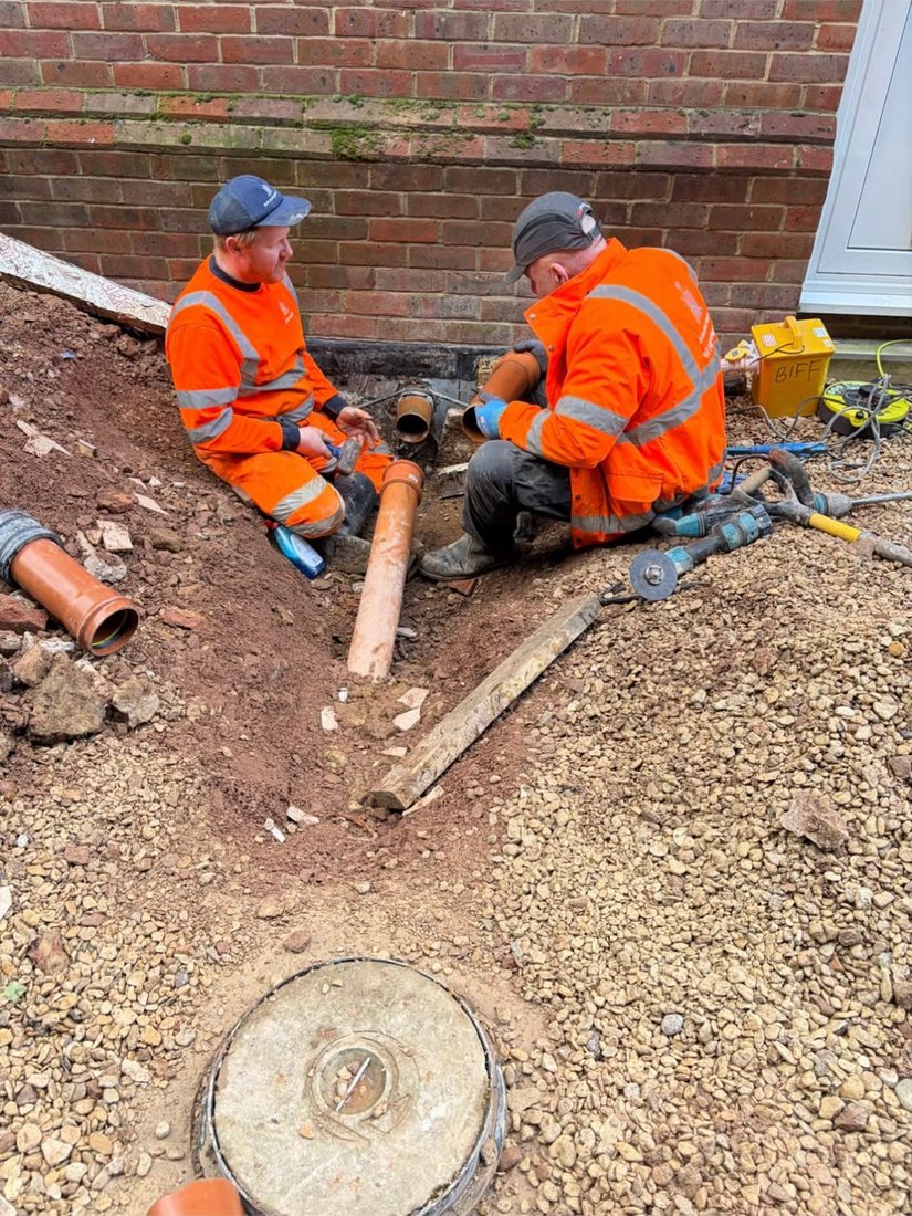 Two DrainAway engineers in orange high-visibility kit in an excavation against a house wall, fitting a new length of pipe.