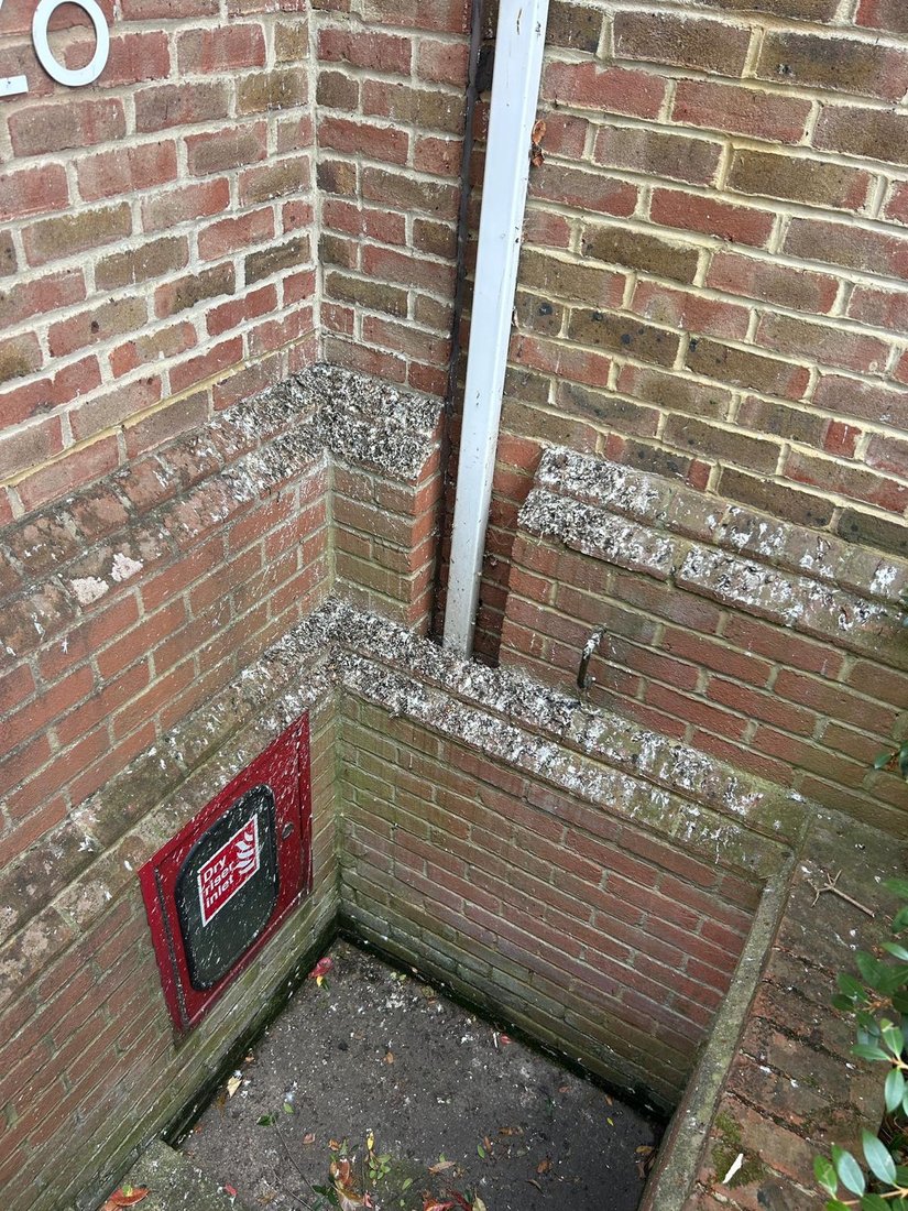 A gully in the corner of a brick service well, with a rainwater pipe running down the wall and a dry riser inlet beside it.