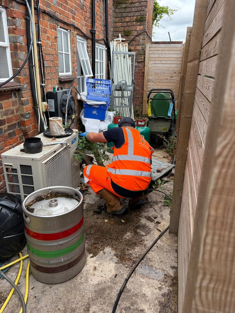 A DrainAway engineer in high-visibility kit working at an open chamber in a narrow pub yard, beside beer kegs and air conditioning units.