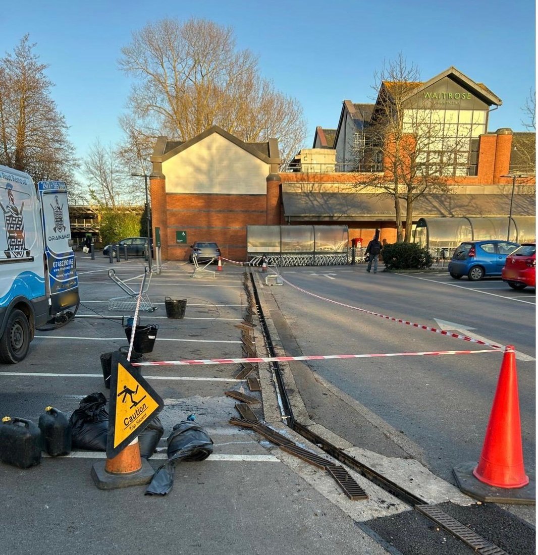 A channel drain lifted for replacement across a supermarket car park, with barrier tape, cones and a DrainAway van set up around the works.