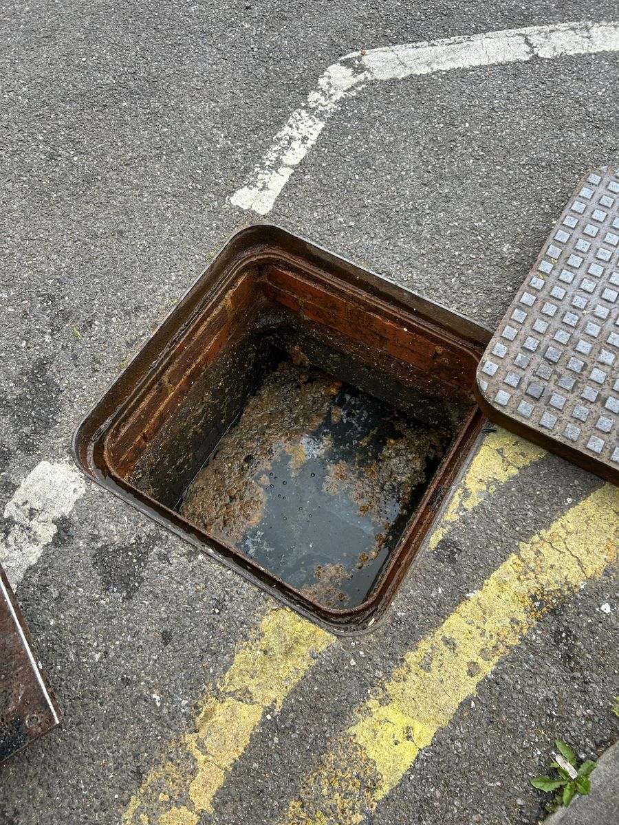 A roadside inspection chamber on double yellow lines, its base holding silt, scum and standing water.