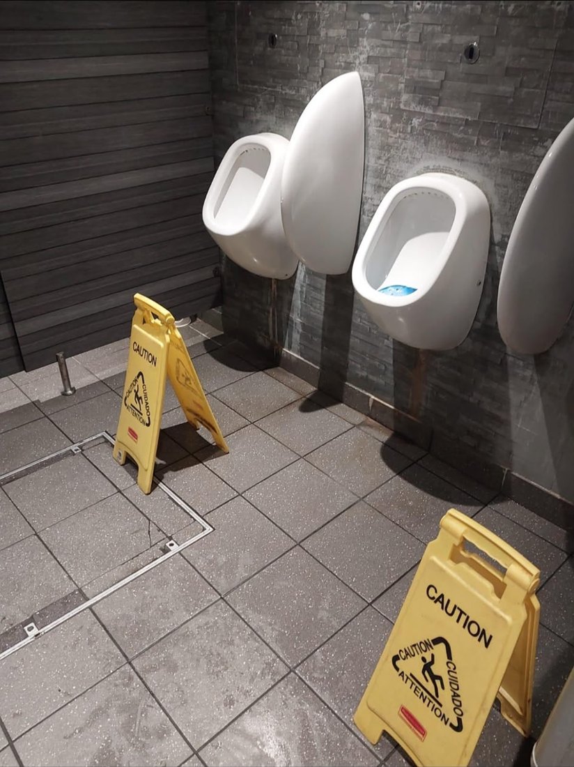 A row of urinals in a commercial washroom with caution wet floor signs out and an access chamber set into the tiled floor.