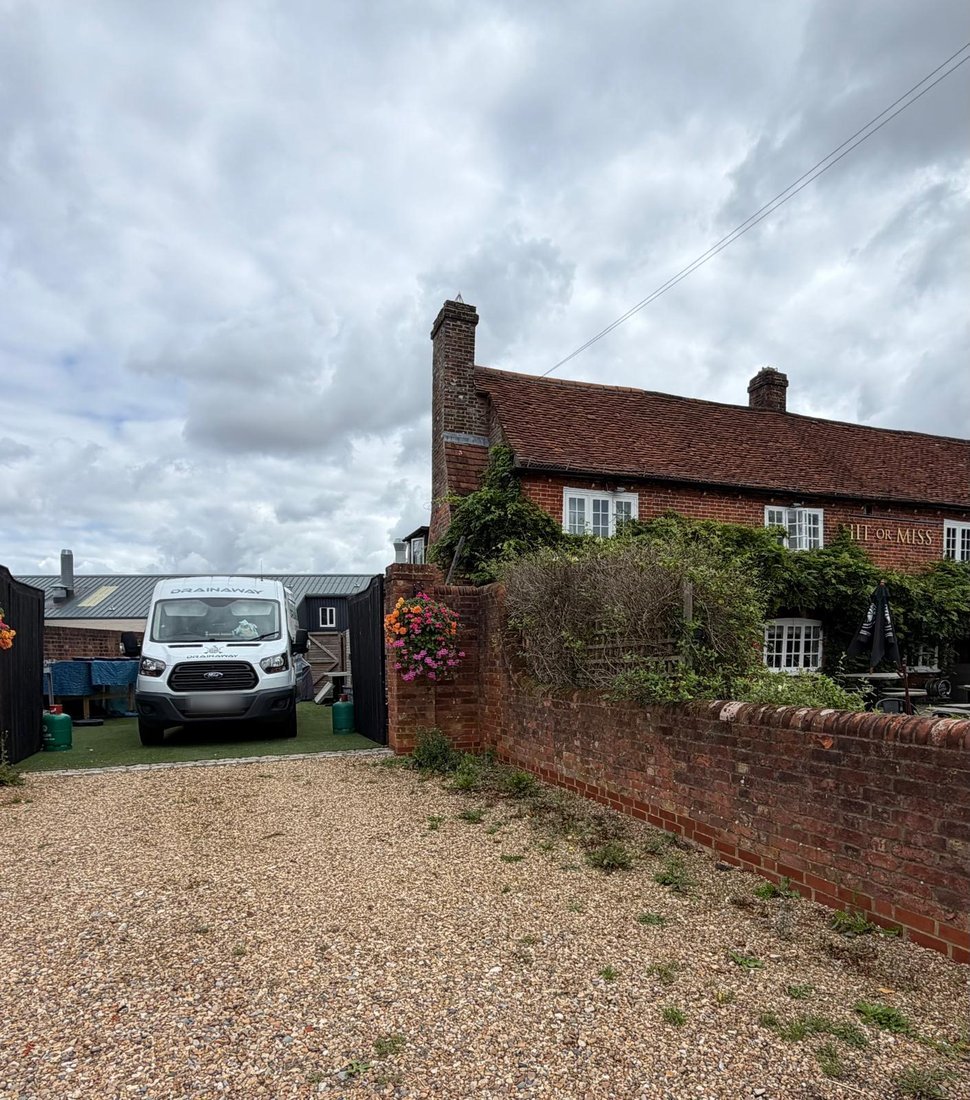 A DrainAway van parked in the yard of a village pub, with the pub's brick frontage and garden beyond the wall.