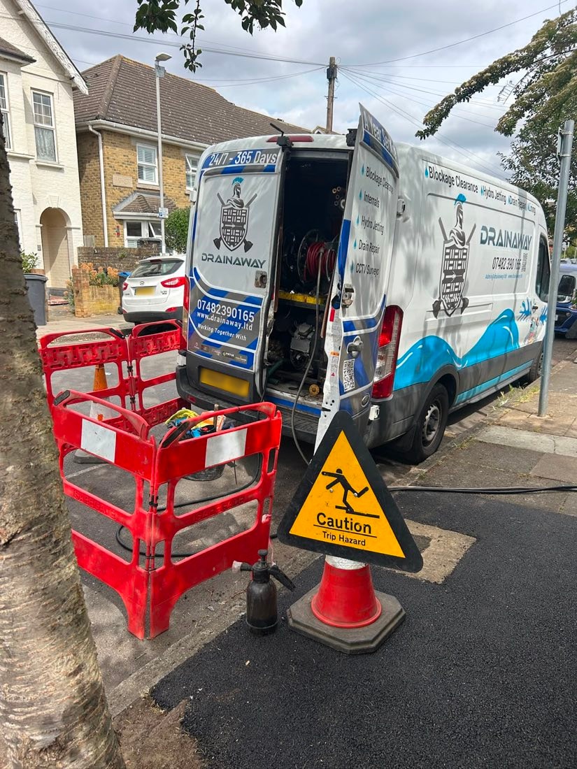 The rear of a DrainAway van open on a residential street showing jetting equipment inside, with barriers and cones set out.