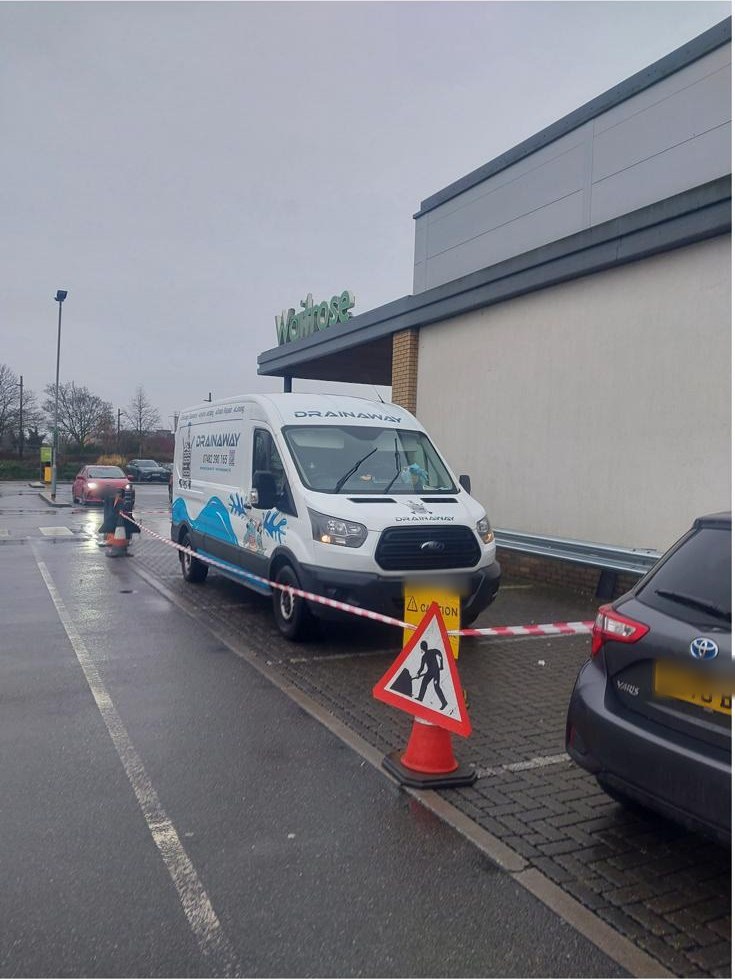A DrainAway van taped off beside a supermarket in the rain, with a cone and a works sign set out around the job.