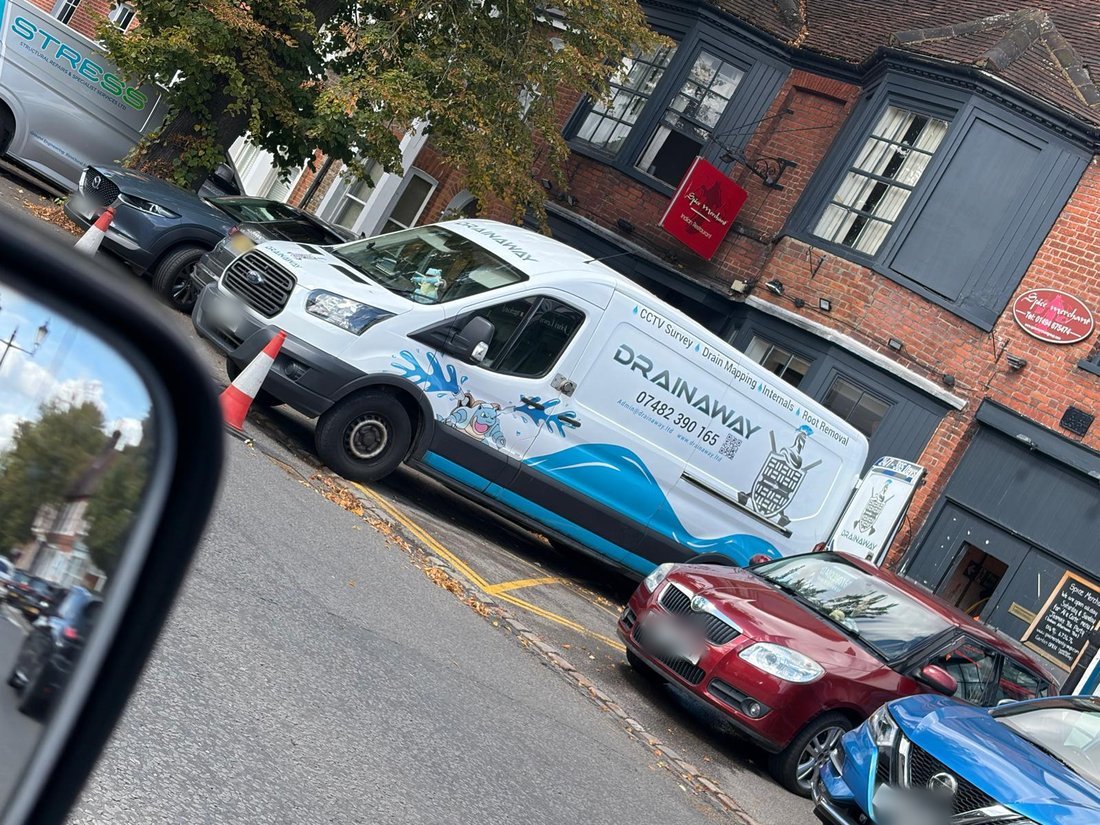A liveried DrainAway van parked on a town-centre street outside a row of shops, with a cone set out at its front.