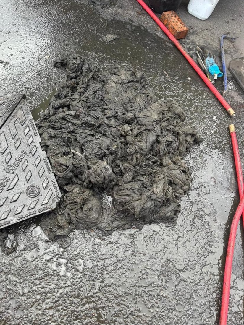 A large mass of wet wipes pulled out of a sewer and piled beside the open chamber cover, with the jetting hose alongside.