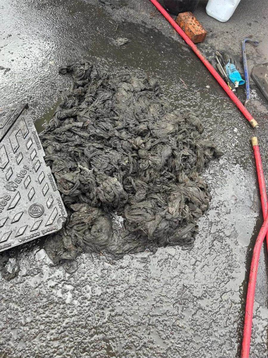 A large mass of grey wipes pulled out of a drain and laid on the wet road beside the lifted cover, with a jetting hose alongside.