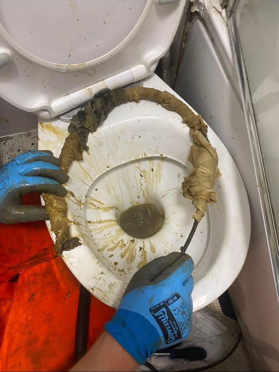 A long rope of wet wipes pulled out of a toilet and draped over the rim, with an engineer working a hand auger in blue gloves.